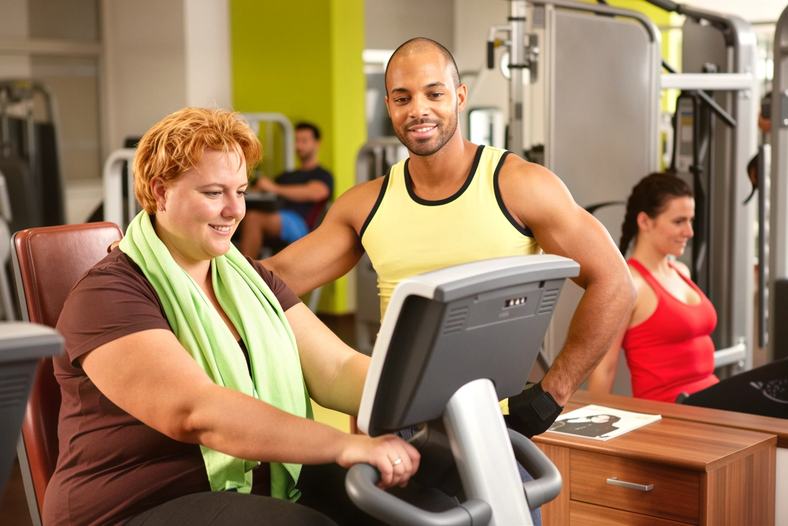 Plus-size woman in athletic wear sitting on a stationary exercise bike with a green towel around her neck, guided by a male personal trainer in a gym studio.