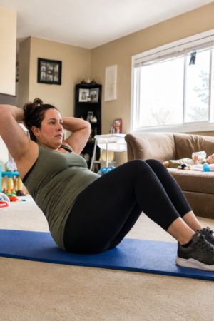 Busy New Mums Mother doing crunches on a blue exercise mat at home while her baby rests on the couch nearby.