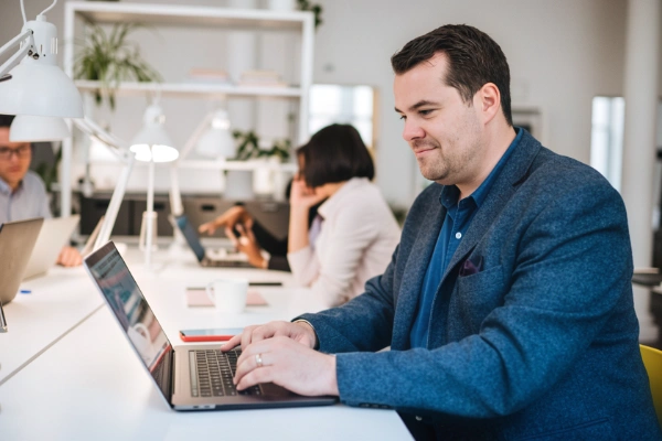 A man in a blue jacket smiling while typing on a laptop computer at a desk in a modern office.