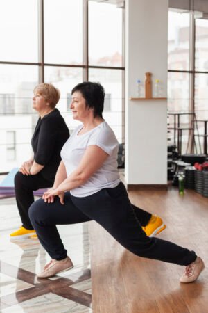 elderly-women-stretching Two senior women performing standing side lunge stretches together in a bright gym studio.