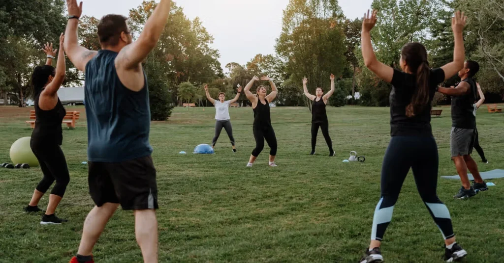 Diverse group of people performing jumping jacks together during an outdoor group fitness workout in a grassy park.