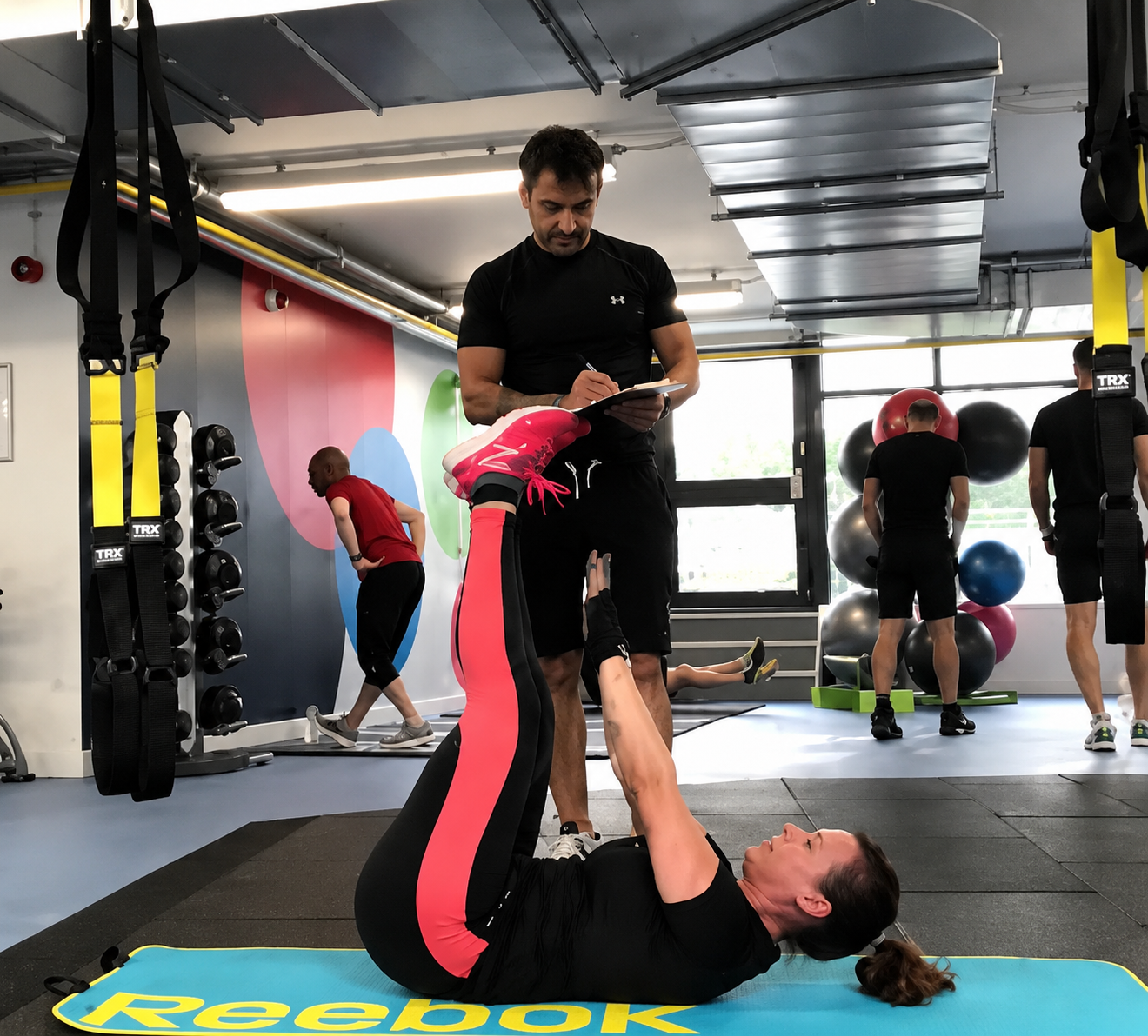 Woman lying on a fitness mat performing vertical leg raises while a male personal trainer takes notes on a clipboard in a gym.