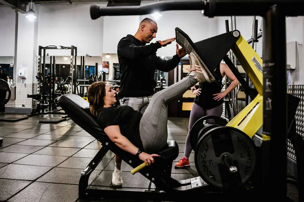 A woman performing a leg press exercise on a gym machine while a male personal trainer stands nearby guiding her form.