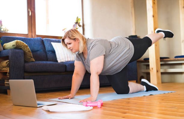 Plus-size woman on hands and knees performing a leg kickback exercise on a yoga mat in her living room while looking at a laptop screen.
