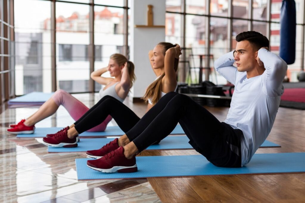A man and two women lined up on blue mats performing sit-ups during a fitness class in a bright gym studio.