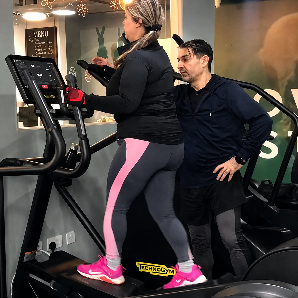 Woman exercising on a Technogym stair climber machine while a male personal trainer stands beside her guiding her form.