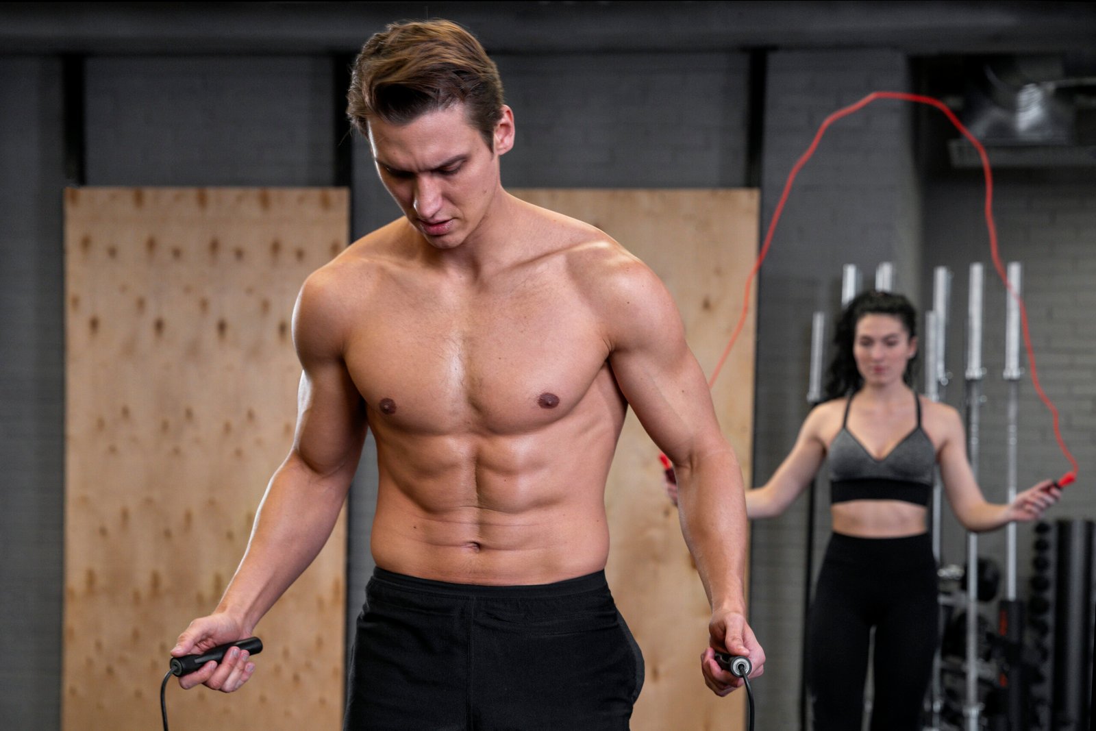 Shirtless athletic man jumping rope in the foreground while a woman skips rope behind him in a gym.