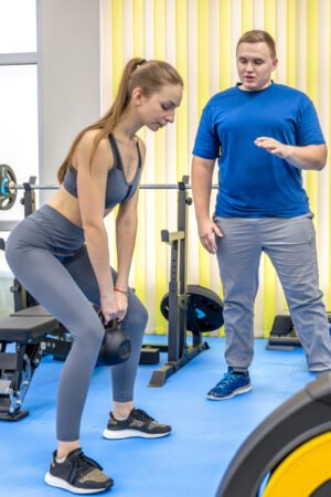 two-people-rehab-doctor-teaches-woman-kettlebell-deadlift-movement-control Woman performing a kettlebell deadlift while a male personal trainer coaches her form in a gym.